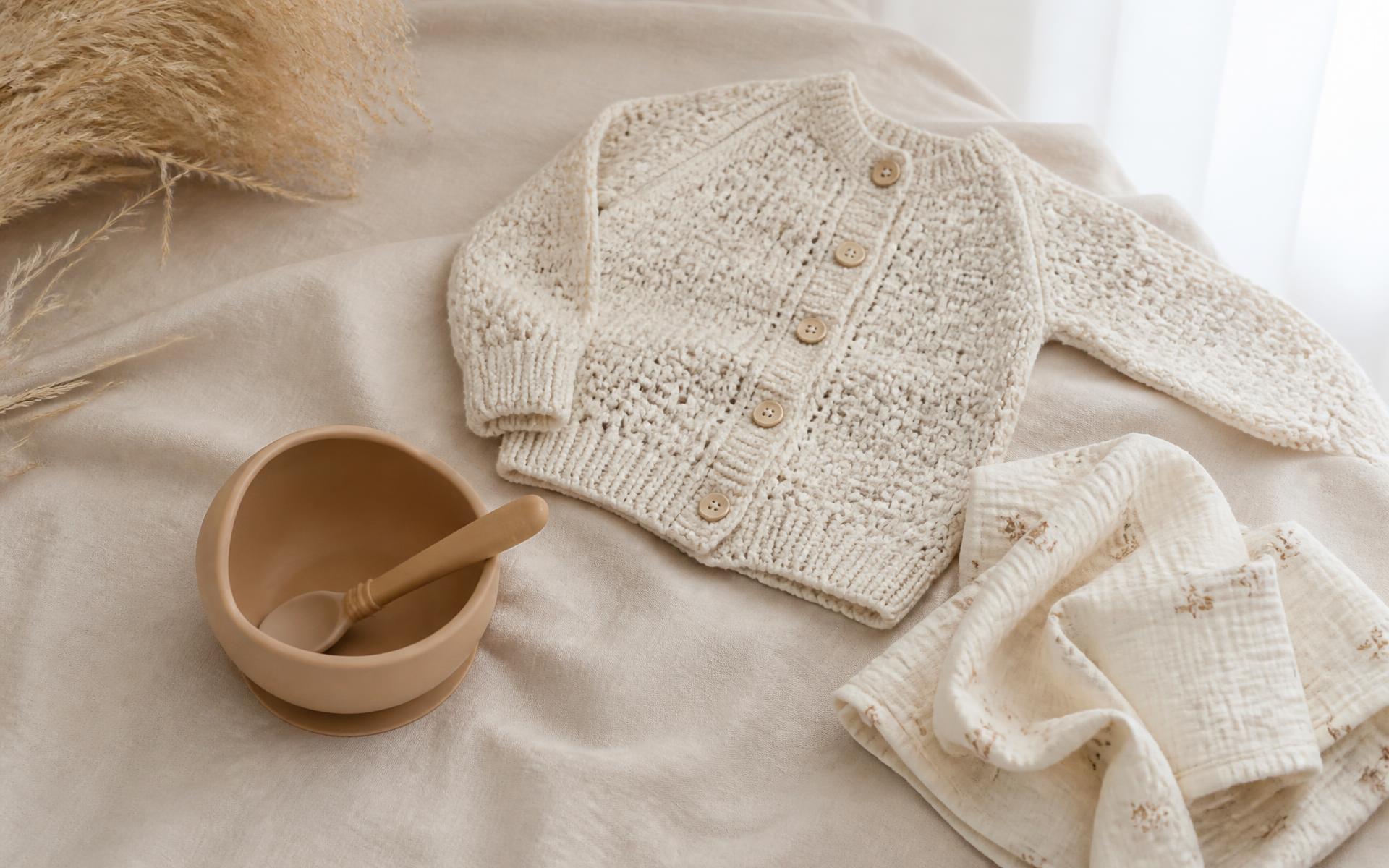 Baby cardigan, silicone feeding bowl and muslin cloths on warm linen